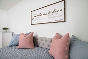 Un dormitorio con cama, dos almohadas, una mesita de noche con planta y una cita enmarcada en la pared.