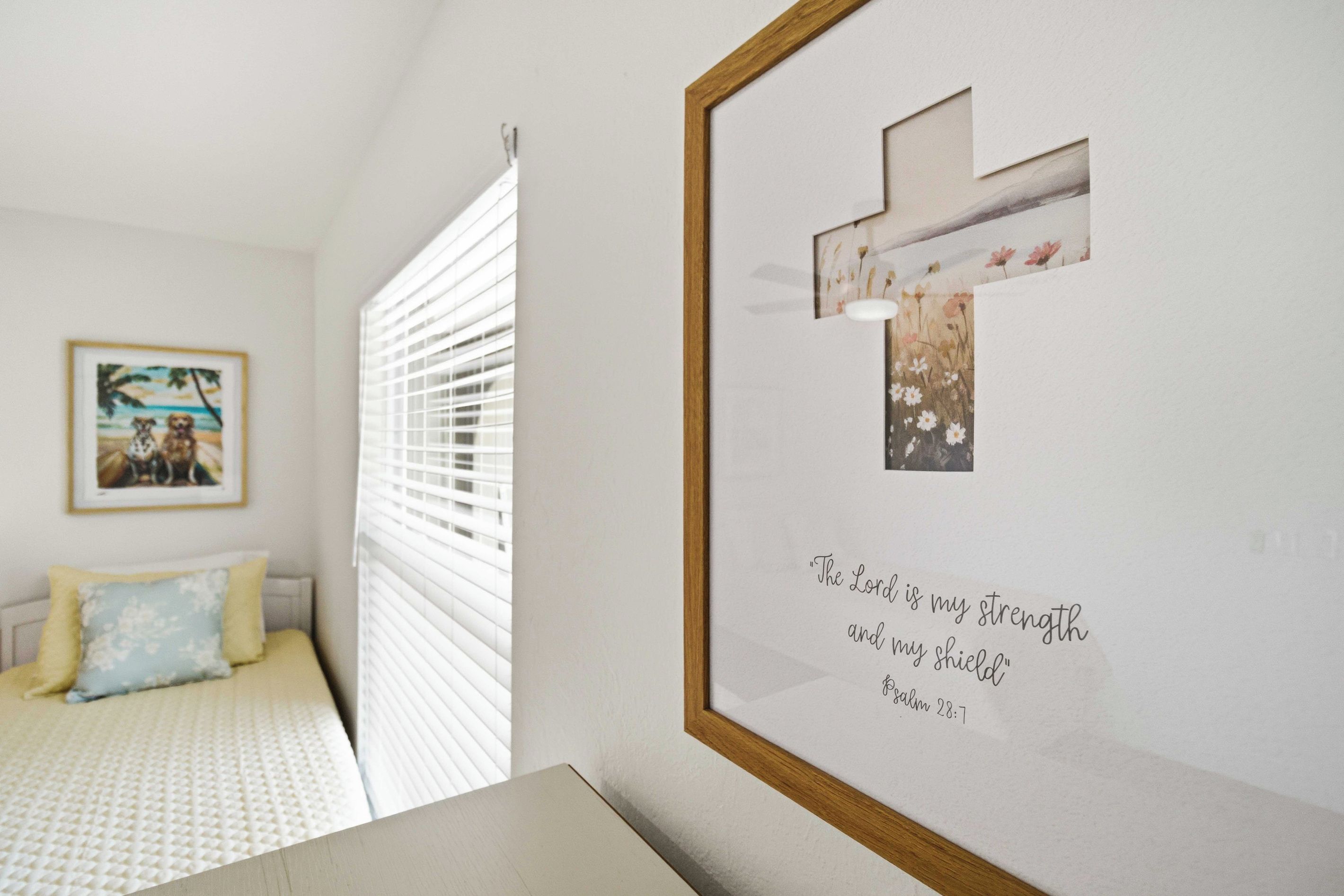 A bedroom with a bed, a framed religious quote, and a window with blinds.