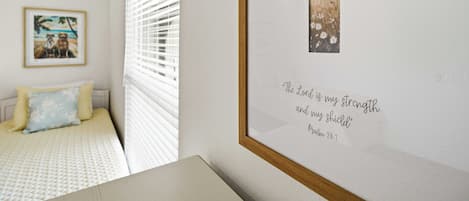 A bedroom with a bed, a framed religious quote, and a window with blinds.