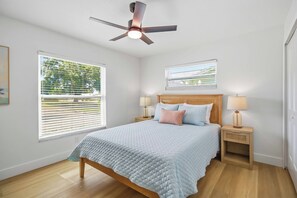 Une chambre à coucher comprenant un lit, deux tables de chevet avec des lampes, un ventilateur de plafond et deux fenêtres équipées de stores.