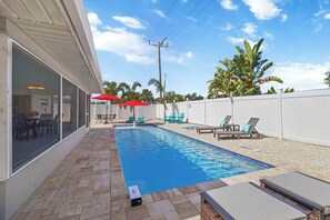 A swimming pool with lounge chairs and umbrellas, surrounded by a white fence and palm trees. - Newly Remodeled w/ Heated Pool! Near Anna Maria! (Bradenton)