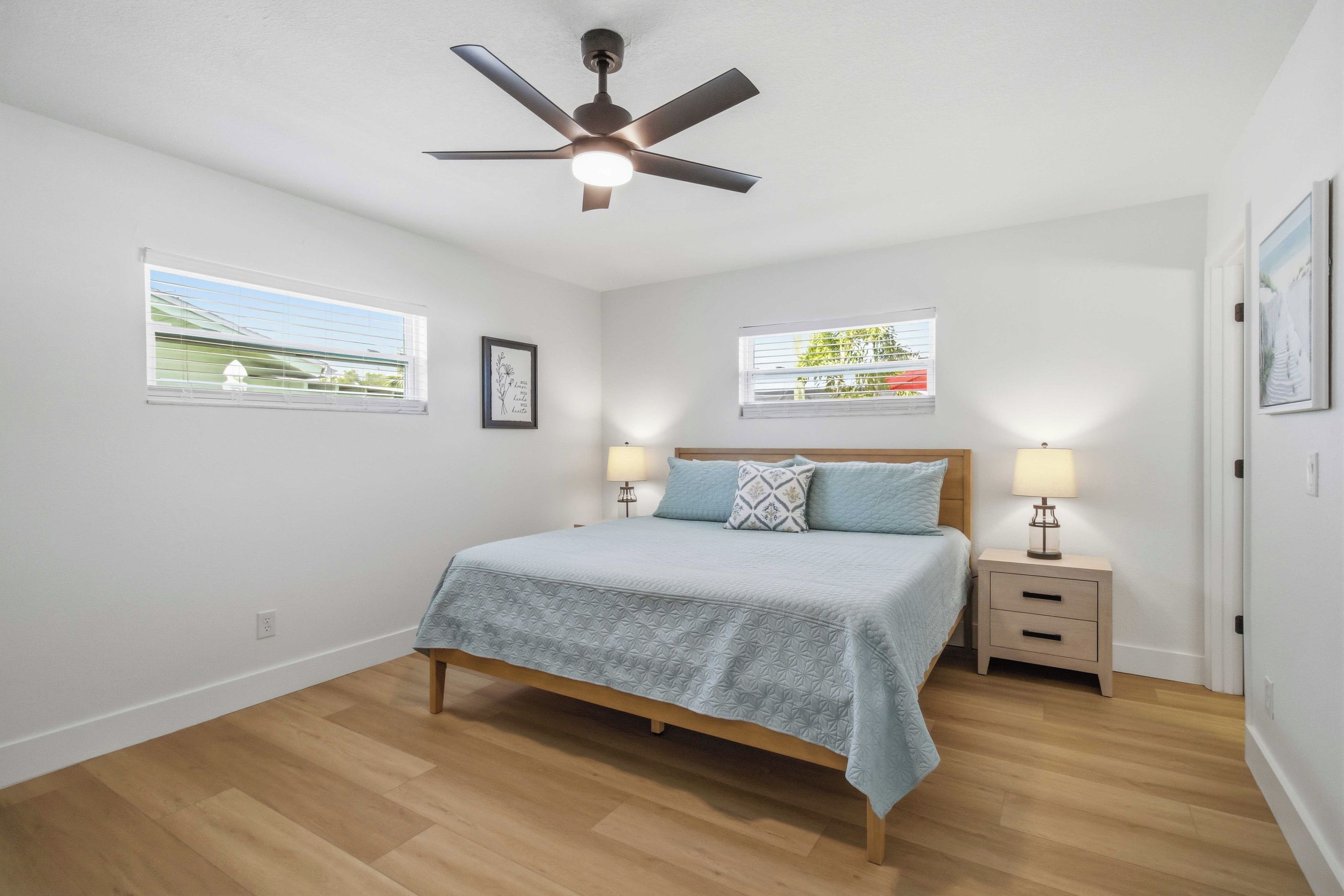 Une chambre à coucher comprenant un lit, deux tables de chevet avec des lampes, un ventilateur de plafond et deux fenêtres équipées de stores.