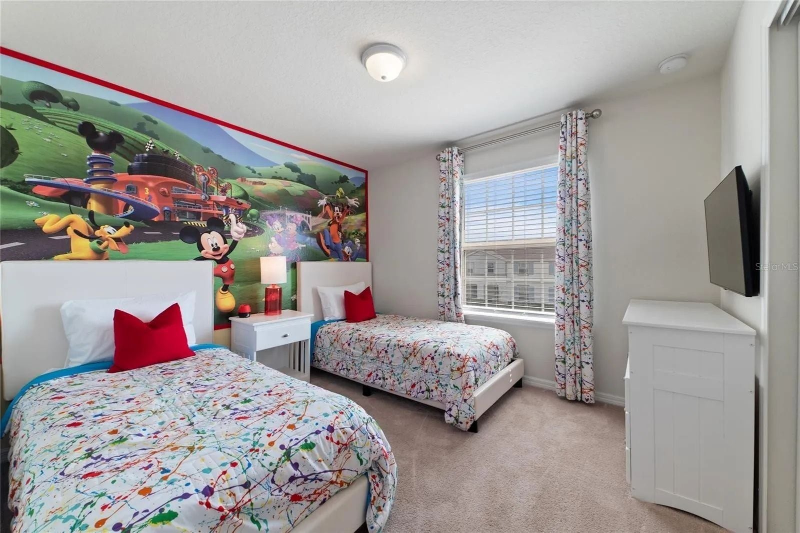 Habitación de hotel con dos camas, un mural grande de Mickey Mouse, una ventana con cortinas y un televisor de pantalla plana montado en la pared.