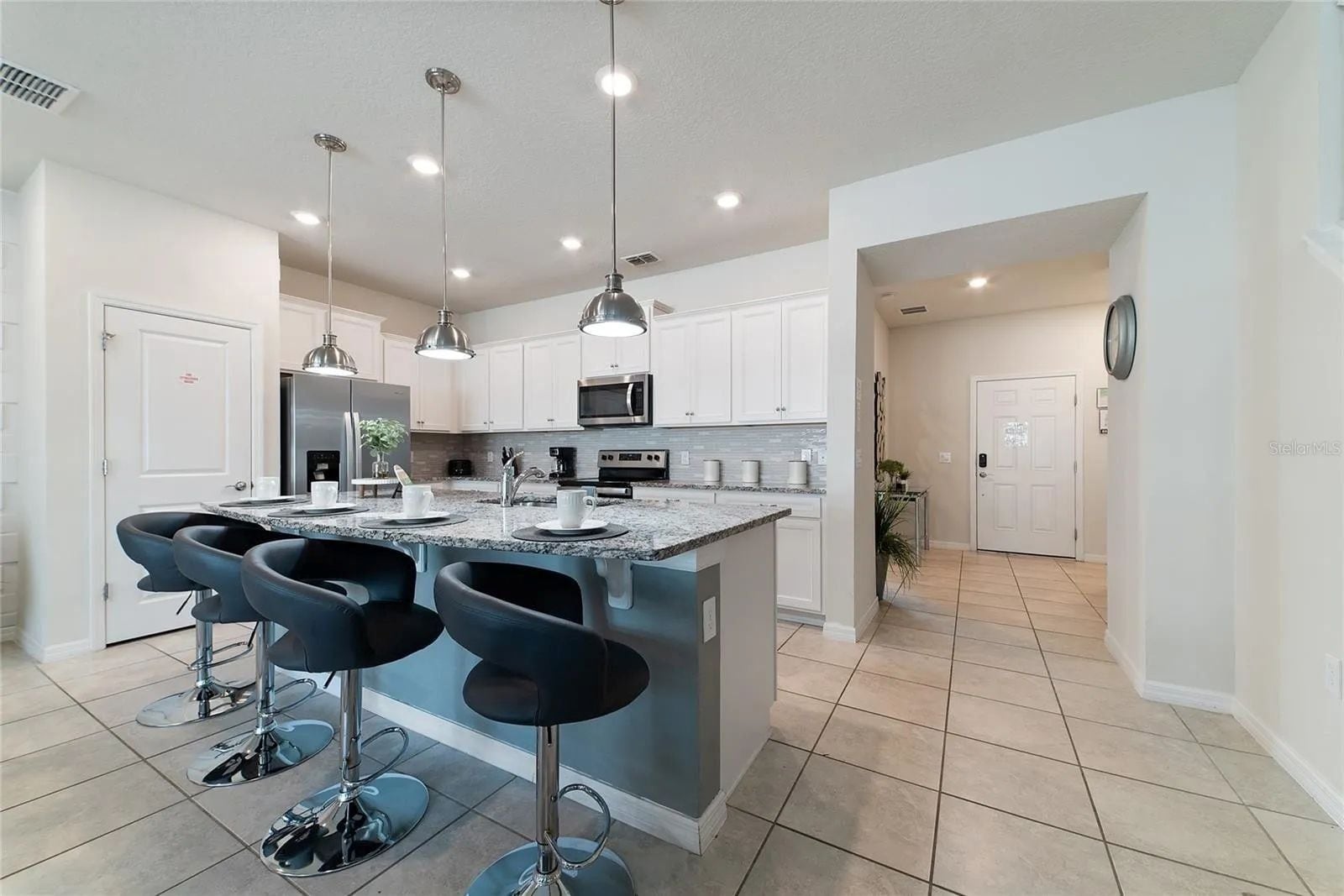 A modern kitchen with a central island, bar stools, stainless steel appliances, pendant lighting, and a tiled floor.