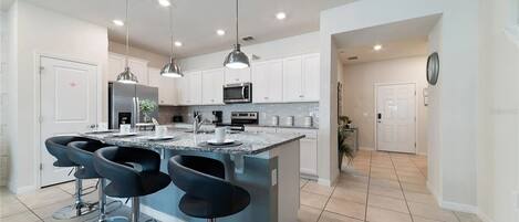 A modern kitchen with a central island, bar stools, stainless steel appliances, pendant lighting, and a tiled floor.