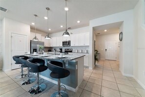 A modern kitchen with a central island, bar stools, stainless steel appliances, pendant lighting, and a tiled floor. - Windsor Island 5BR Home Near Disney Parks (Davenport)