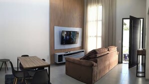 Ein modernes Wohnzimmer mit Fernseher, braunem Sofa, Esstisch mit Stühlen und einem großen Fenster mit Vorhängen.