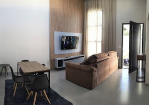 Un salon moderne doté d’un téléviseur à écran plat, d’un canapé marron, d’une table à manger avec des chaises et d’une grande fenêtre agrémentée de rideaux.