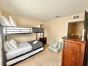 Llits superposats amb roba de llit blanca, una petita taula de nit de fusta, una banca amb coixí verd i un armari de fusta amb una televisió.