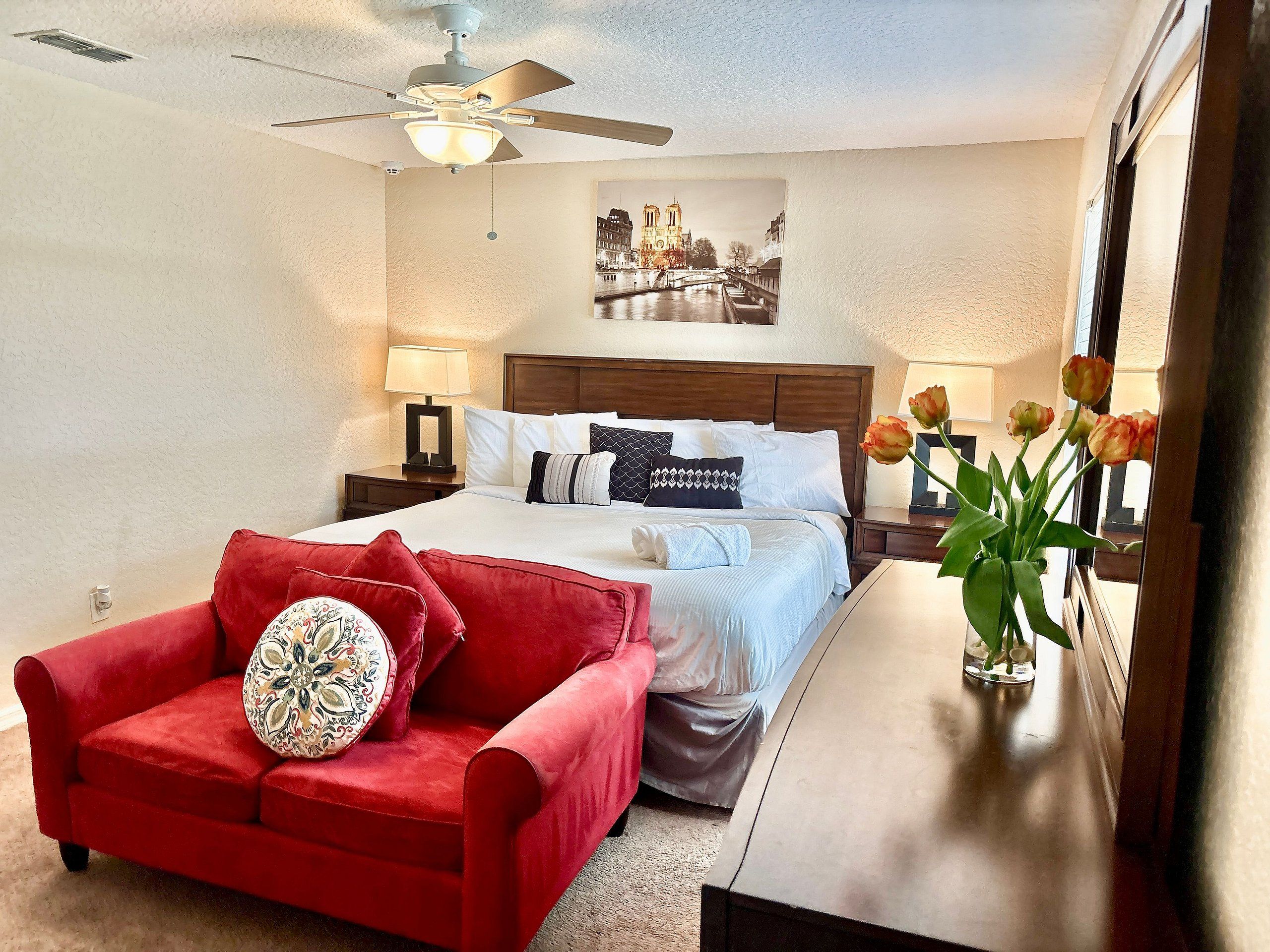 Habitación de hotel con cama, sofá rojo, mesita de noche con lámpara, ventilador de techo y ventana con vista a un puente.