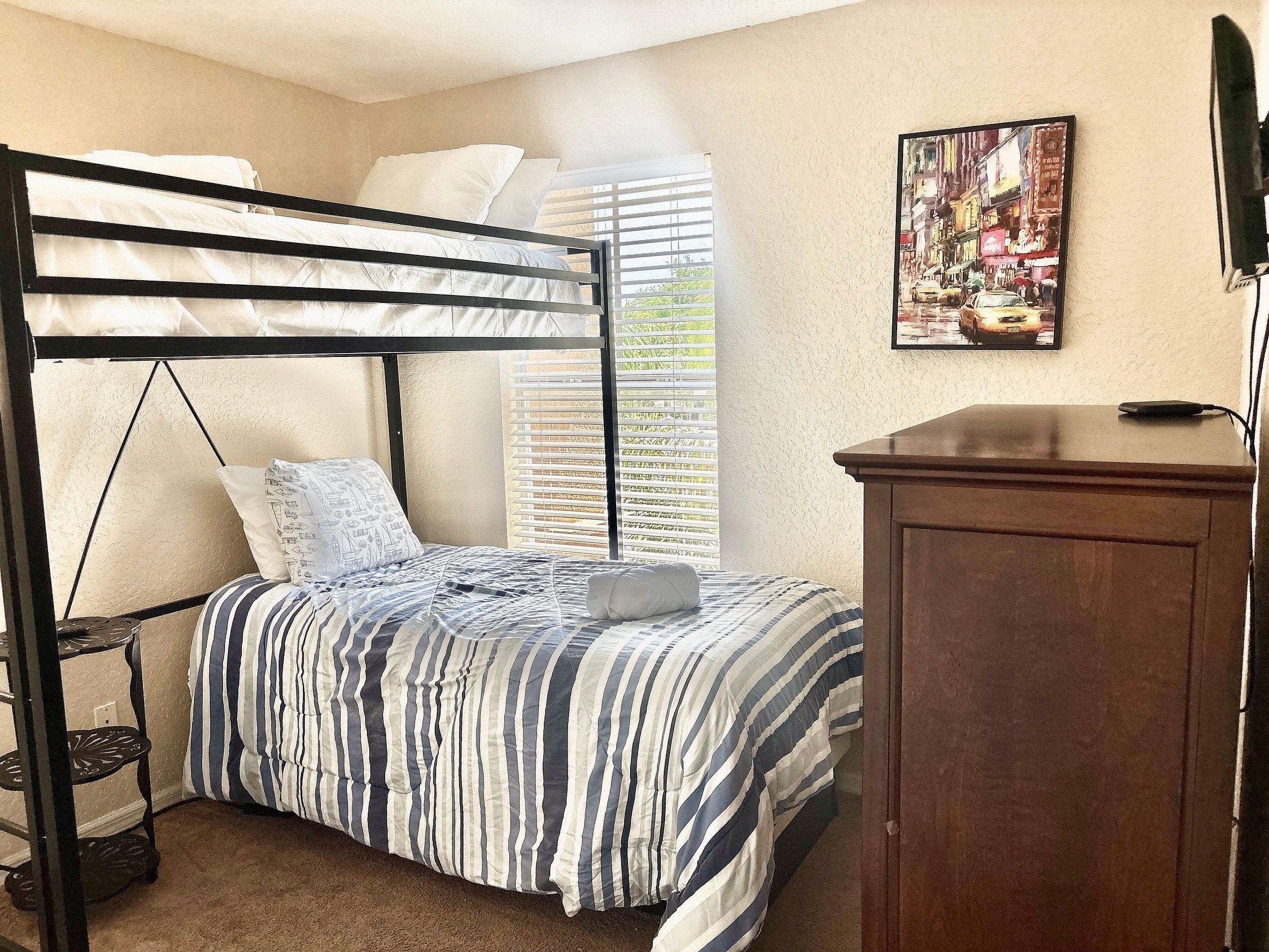 A bunk bed with a striped bedspread, a wooden cabinet, and a window with blinds.