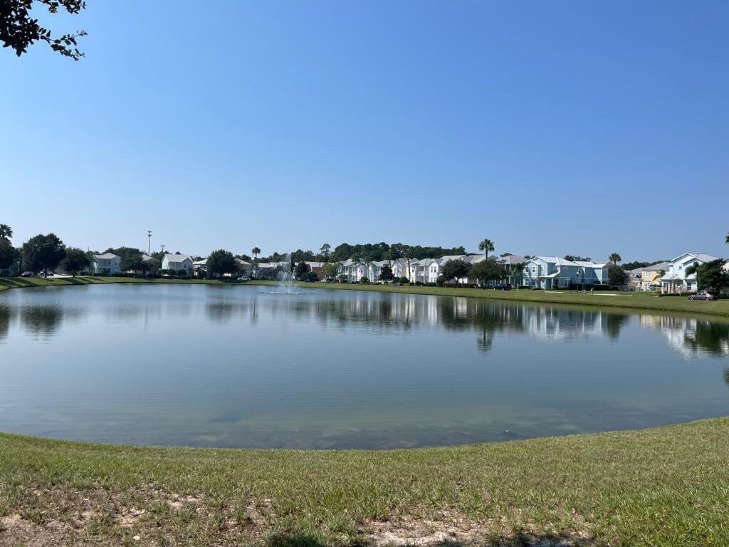 Un lac paisible avec une fontaine au centre, entouré d’une zone herbeuse et de bâtiments résidentiels en arrière-plan.