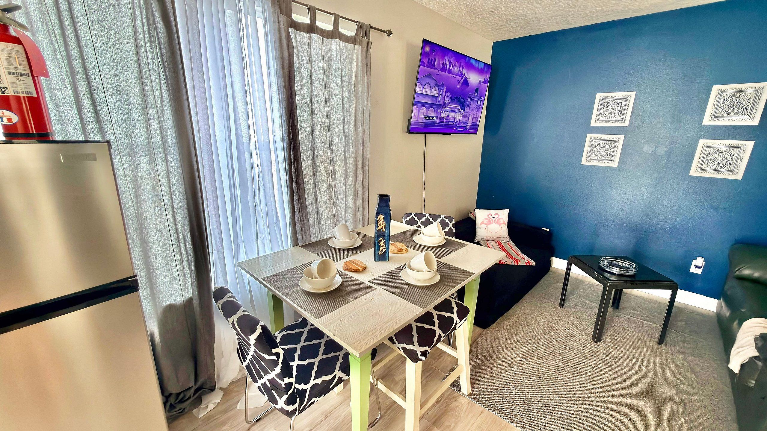A compact living space with a kitchenette, a dining area with a table and chairs, a blue accent wall, and a television mounted above the table.