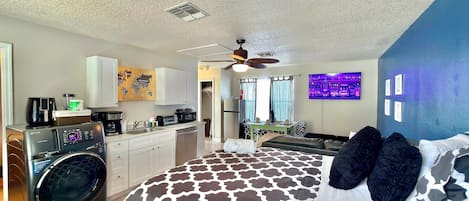 A compact hotel room with a kitchenette, a bed with patterned bedding, a ceiling fan, and a TV mounted on the wall.
