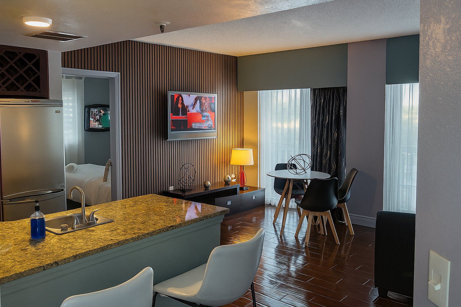 Une chambre d’hôtel équipée d’une kitchenette, d’un coin repas avec une table et des chaises, d’un téléviseur fixé au mur et d’une fenêtre avec des rideaux.