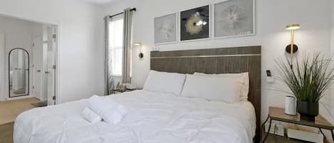 Una cama bien hecha con ropa de cama blanca, una mesita de noche con una lámpara y una planta en maceta.