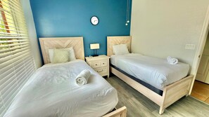 Ein Hotelzimmer mit zwei Betten, einem Nachttisch, einer Wanduhr und einem Fenster mit Jalousien.