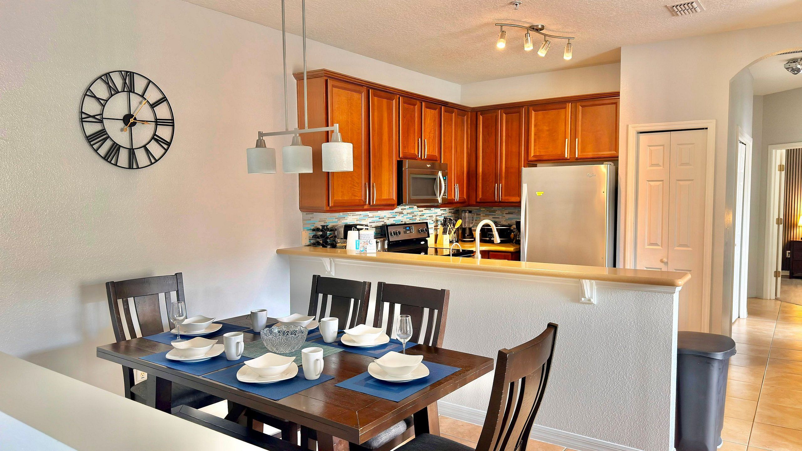 A dining area with a table set for four, a kitchen with wooden cabinets, and a wall clock.