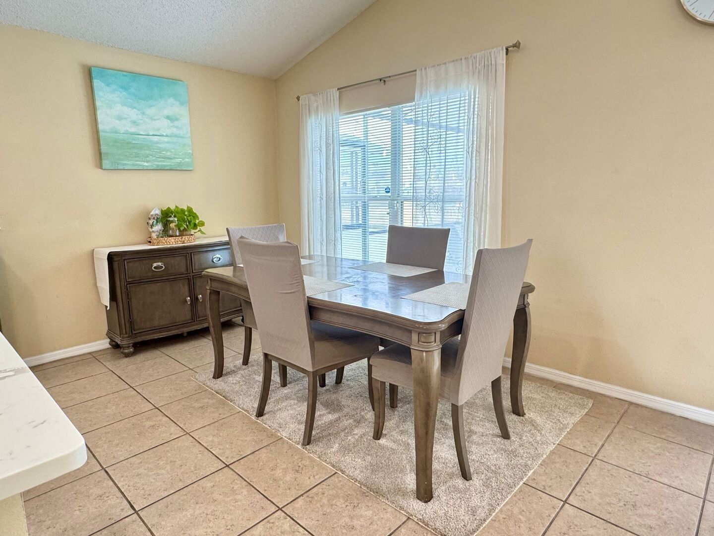 Un coin repas comprenant une table, des chaises, une fenêtre avec des stores, un tableau accroché au mur et un petit meuble contenant des objets décoratifs.