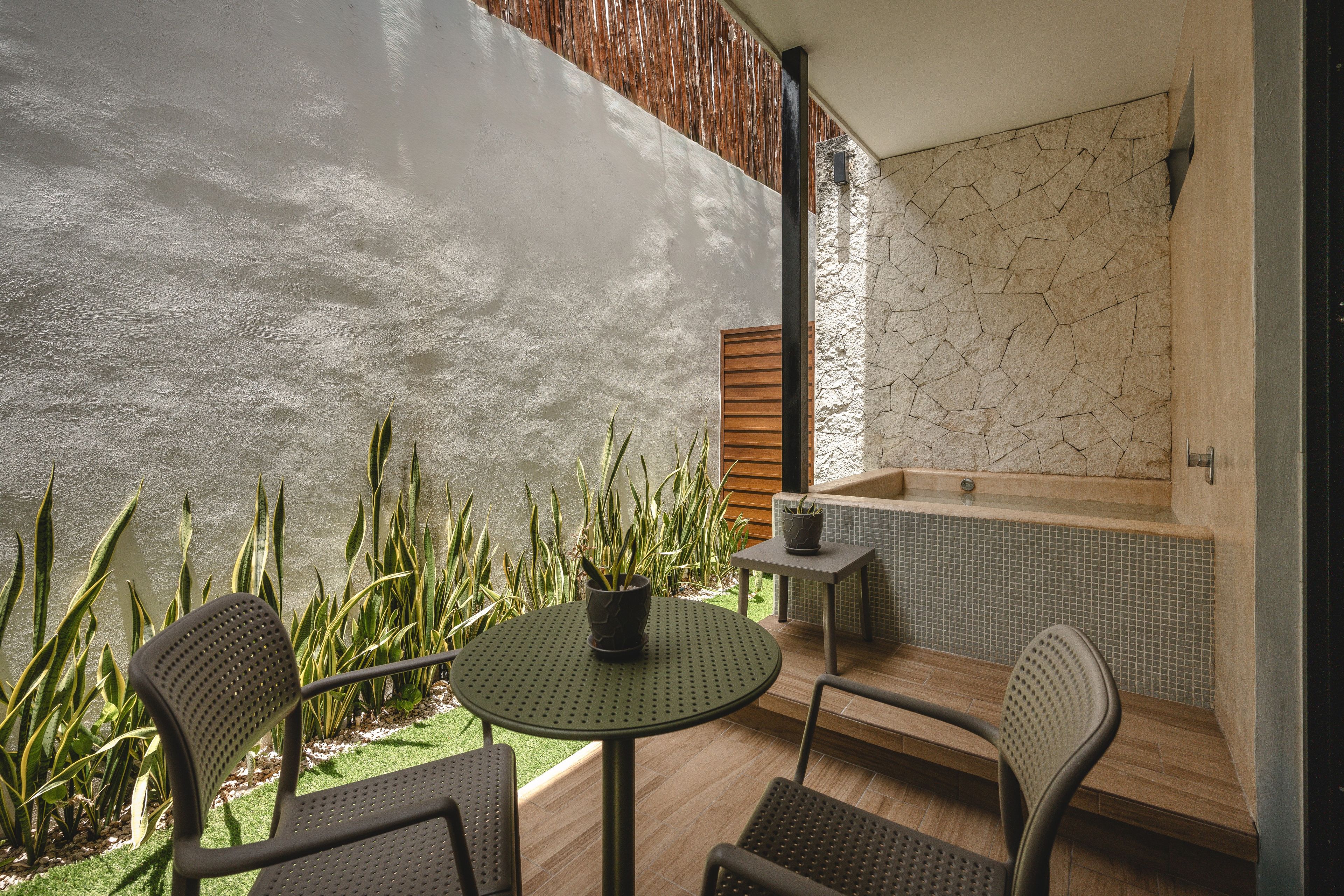 Een patio met een ronde tafel en twee stoelen, een kleine potplant en uitzicht op een wellnessruimte met een betegelde muur en een houten lambrisering.