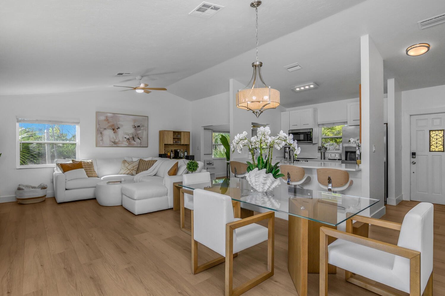 A modern living room with a glass dining table, white sofas, and a ceiling fan.