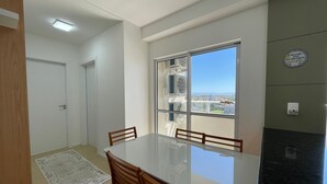Uma sala de jantar com mesa e cadeiras, janela com aparelhos de ar condicionado e vista para edifícios.