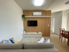 Uma sala de estar moderna com sofá, TV de tela plana fixada em uma parede de madeira, área de jantar com mesa e cadeiras, e um corredor que dá para outro cômodo.