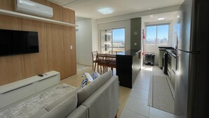 Uma sala de estar moderna com televisão de tela plana, um sofá, área de jantar com cadeiras e cozinha equipada com geladeira e fogão.