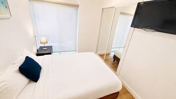 Quarto de hotel com cama, mesa de cabeceira, luminária, televisão e vista para o exterior pela janela.