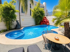 Ein Swimmingpool mit Springbrunnen, umgeben von tropischer Vegetation, einem Tisch mit Stühlen und Blick auf ein Gebäude mit der Aufschrift 'MSI'.