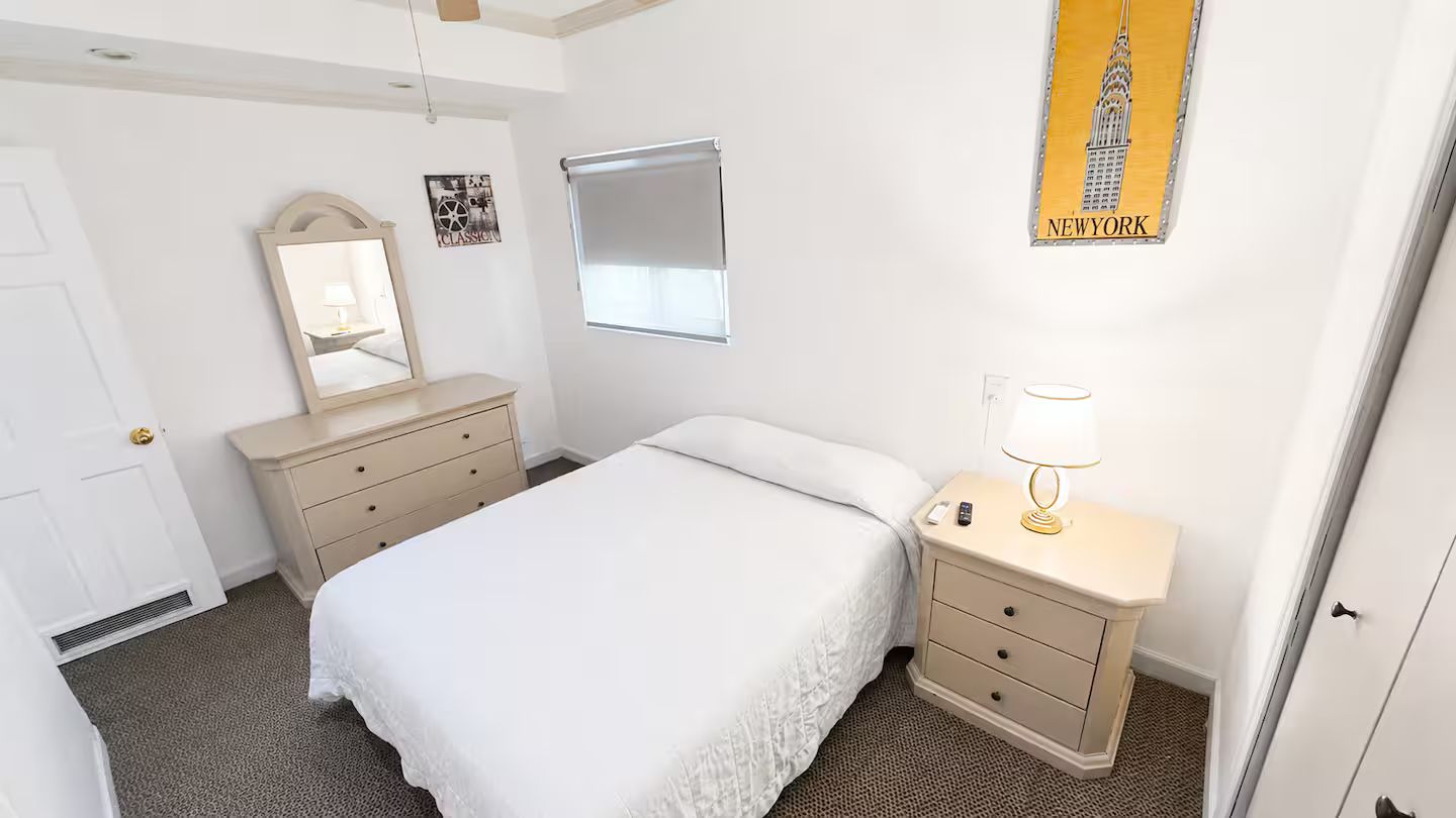 Een hotelkamer met een bed, nachtkastje met lamp, kapstok, raam met blinds en een poster van het Empire State Building.