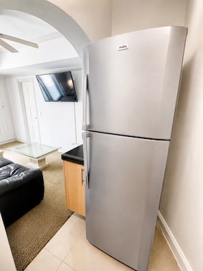A modern kitchen with a stainless steel refrigerator, a glass-top dining table, and a flat-screen TV mounted on the wall.