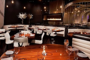 L’intérieur d’un restaurant avec des banquettes, des tables rondes et une cheminée centrale.