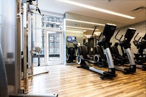 Ein Fitnessraum mit Cardiogeräten, einer Spiegelwand und Blick auf eine Stadtlandschaft durch eine Glastür.