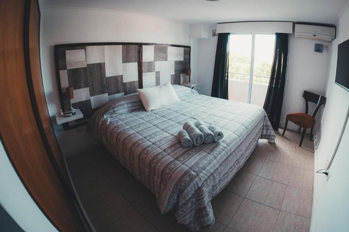 Habitación de hotel con una cama grande, cabecera con diseño geométrico, una mesita de noche con lámpara, una silla y un balcón con cortinas.