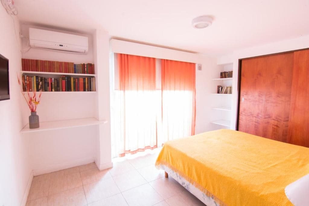 A bedroom with a bed, a bookshelf, a window with curtains, and an air conditioning unit.