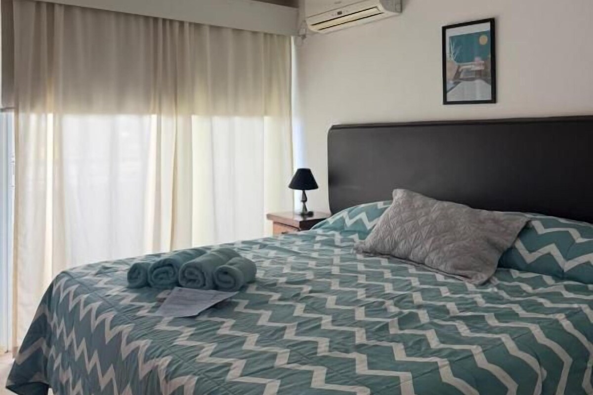 Una habitación de hotel con cama, cortinas, unidad de aire acondicionado, mesita de noche con lámpara y un cuadro en la pared.