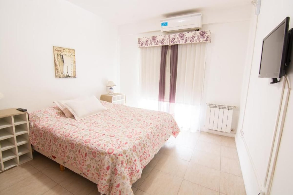 Un dormitorio con una cama, una mesita de noche, una lámpara, un radiador y un televisor fijado a la pared.