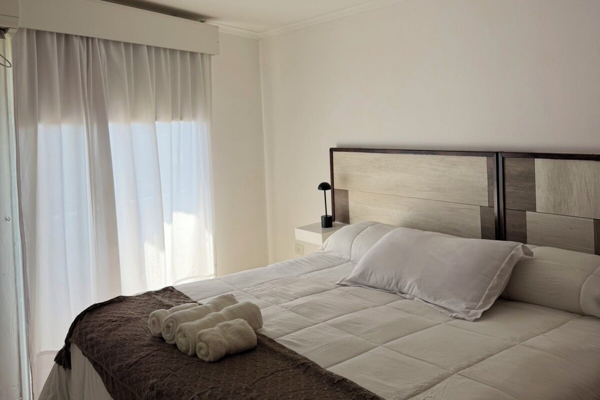 Una habitación de hotel con cama, mesita de noche, lámpara y ventana con cortinas.