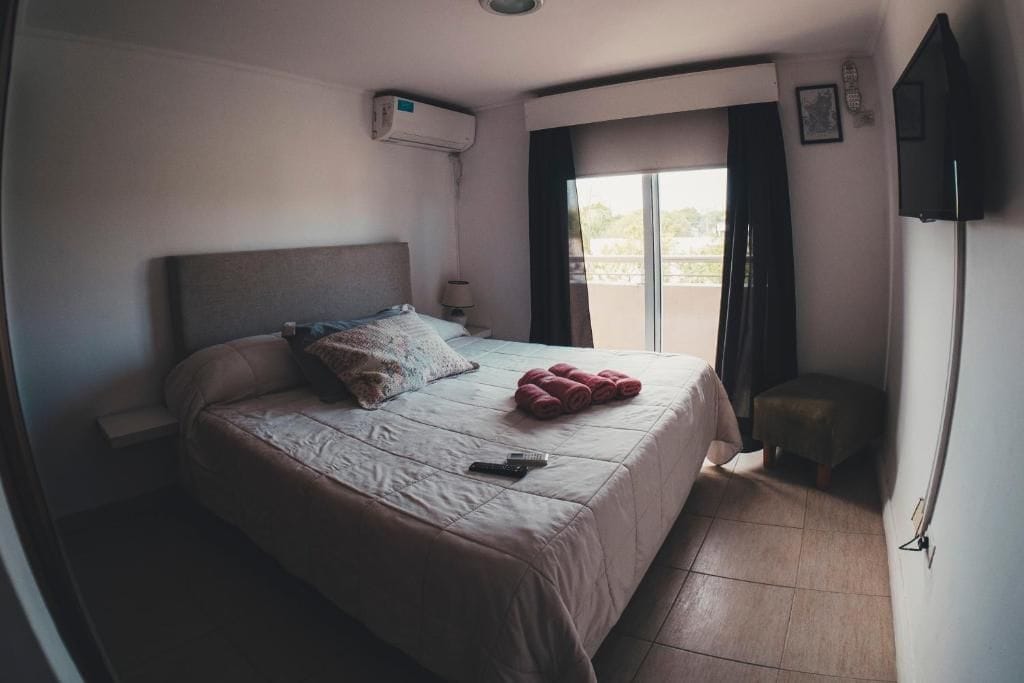 Une chambre d’hôtel comprenant un lit, une petite table, une chaise, un appareil de climatisation, une fenêtre avec des rideaux et un téléviseur à écran plat fixé au mur.