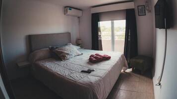 Une chambre d’hôtel comprenant un lit, une petite table, une chaise, un appareil de climatisation, une fenêtre avec des rideaux et un téléviseur à écran plat fixé au mur.