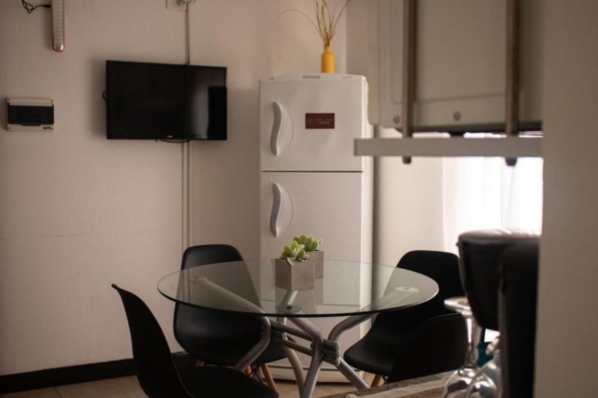 Una habitación moderna con una mesa de vidrio, sillas negras, un refrigerador blanco y un televisor de pantalla plana montado en la pared.