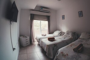 Una habitación de hotel con dos camas, un televisor montado en la pared, una ventana con cortinas y una mesita de noche pequeña.