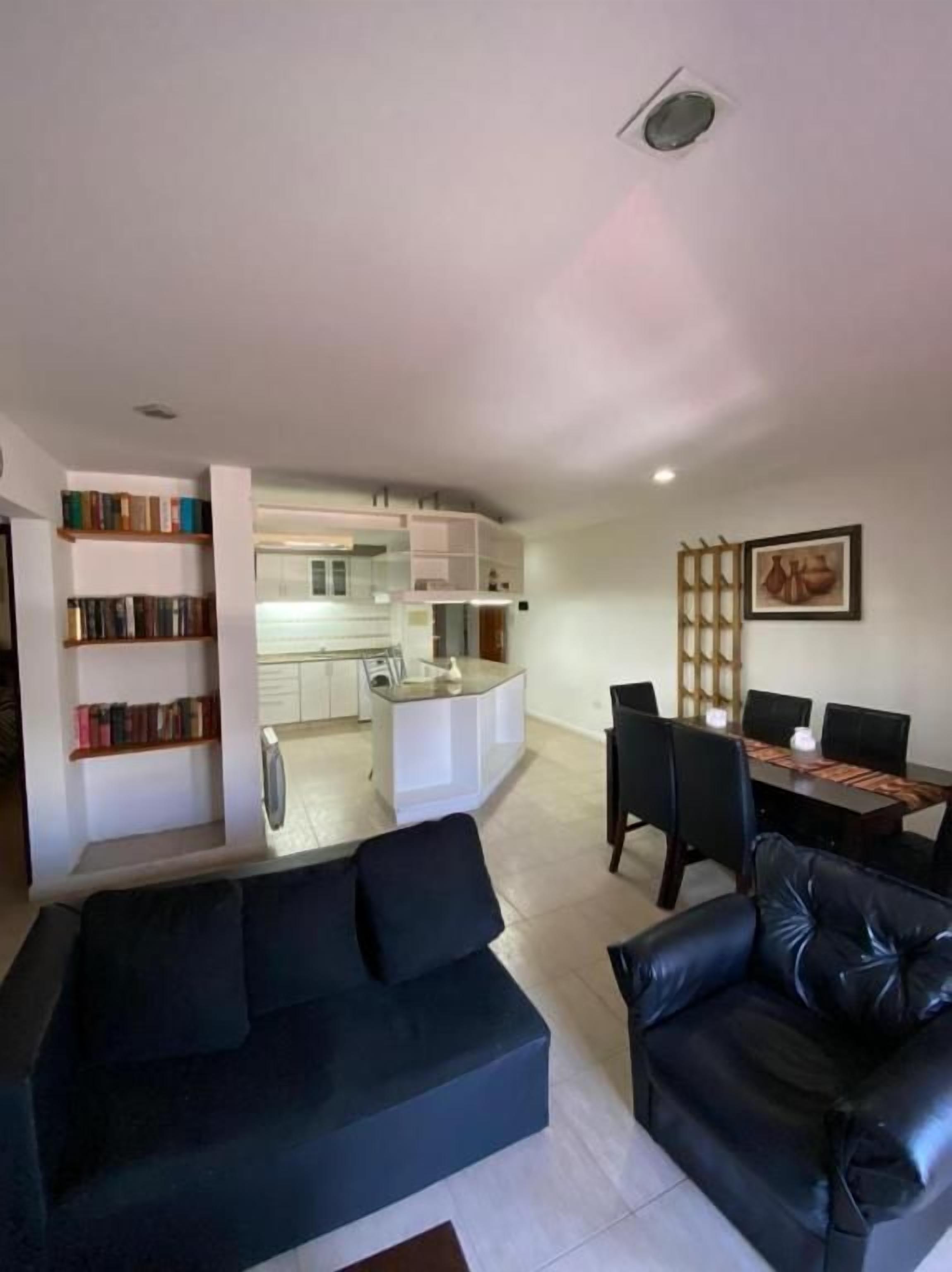 Un salon moderne avec un canapé en cuir noir, un coin repas avec une table et des chaises, et une cuisine en arrière-plan.