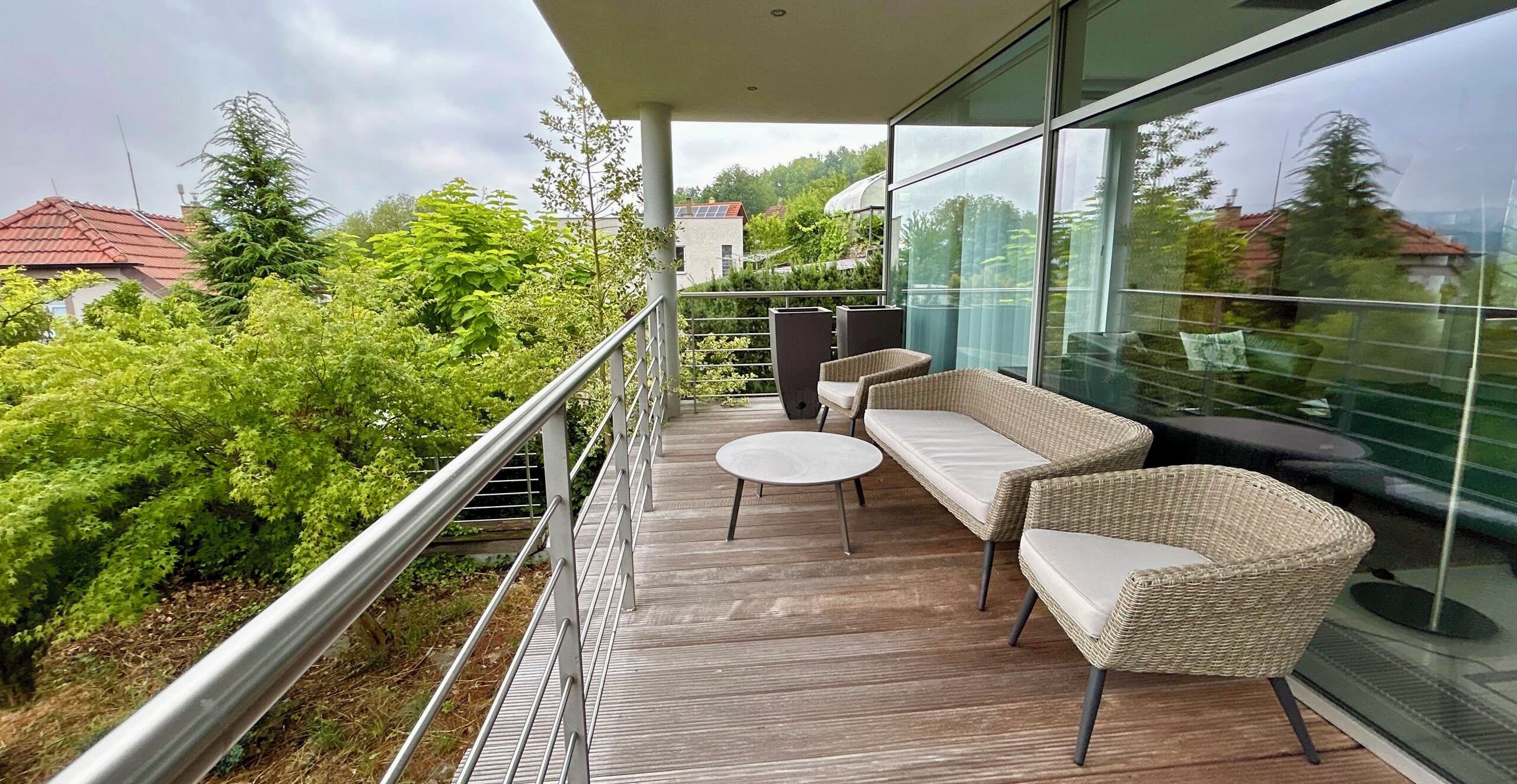 A balcony with a wicker sofa and a small table, overlooking a lush garden and a building with a red roof.