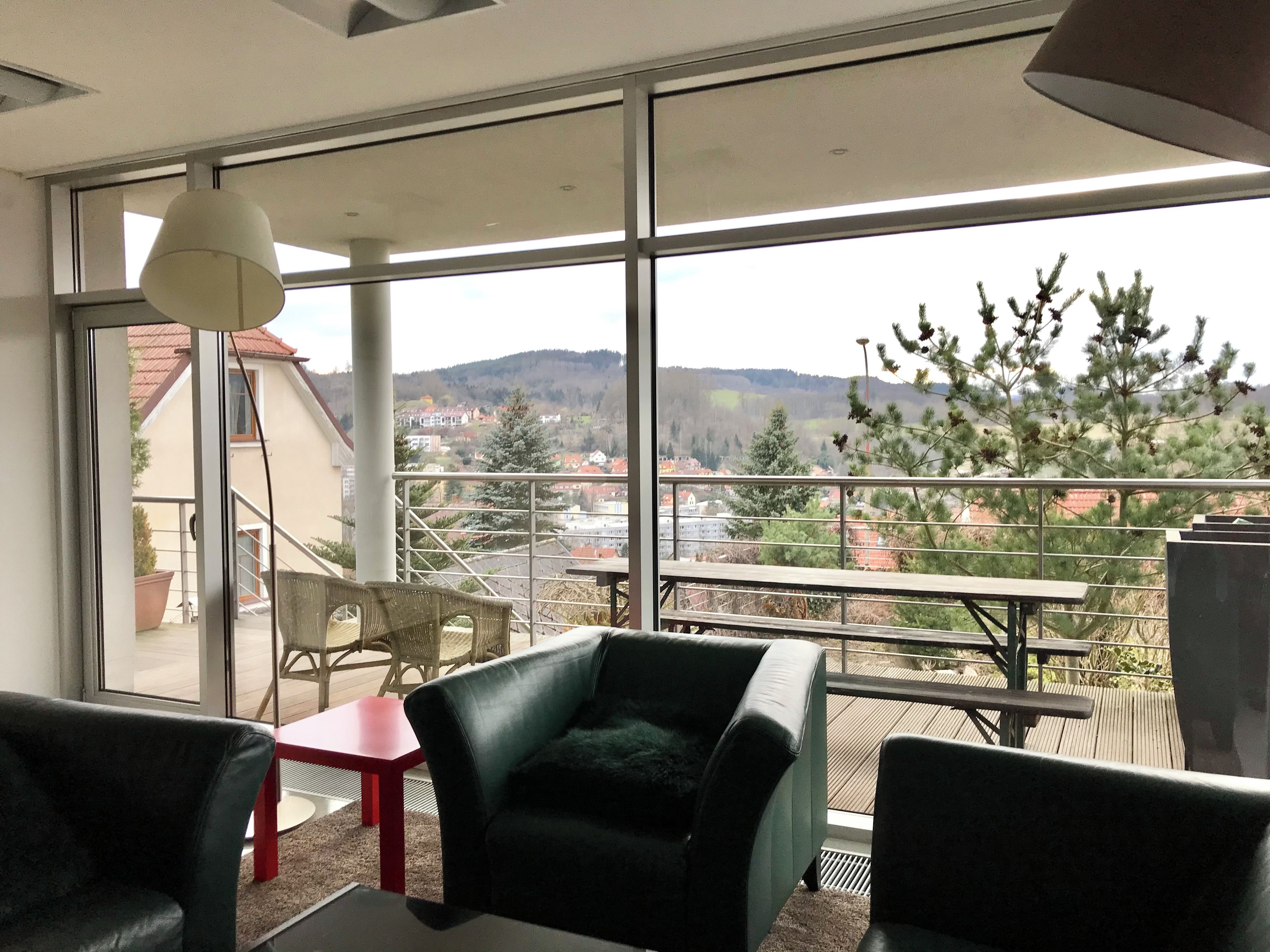 Una habitación moderna con ventanas amplias, un balcón y vistas a un pueblo y colinas.