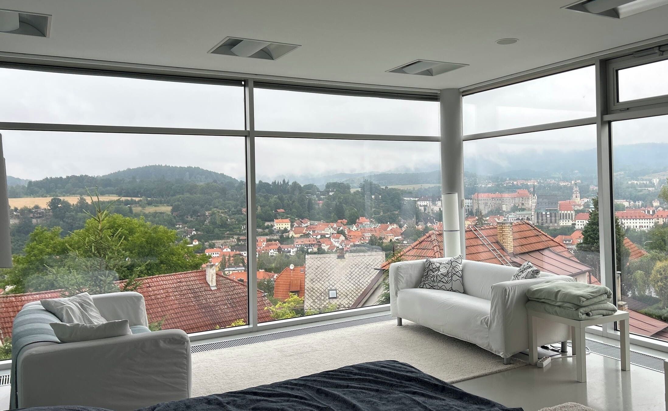 Ein modernes Schlafzimmer mit großen Fenstern, einem Bett, einer Couch und Blick auf eine Stadtlandschaft.