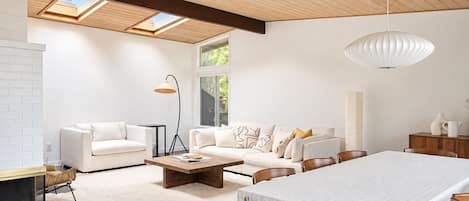 Een moderne woonkamer met een eethoek, met een groot raam, een plafond met dakramen en een witte open haard.