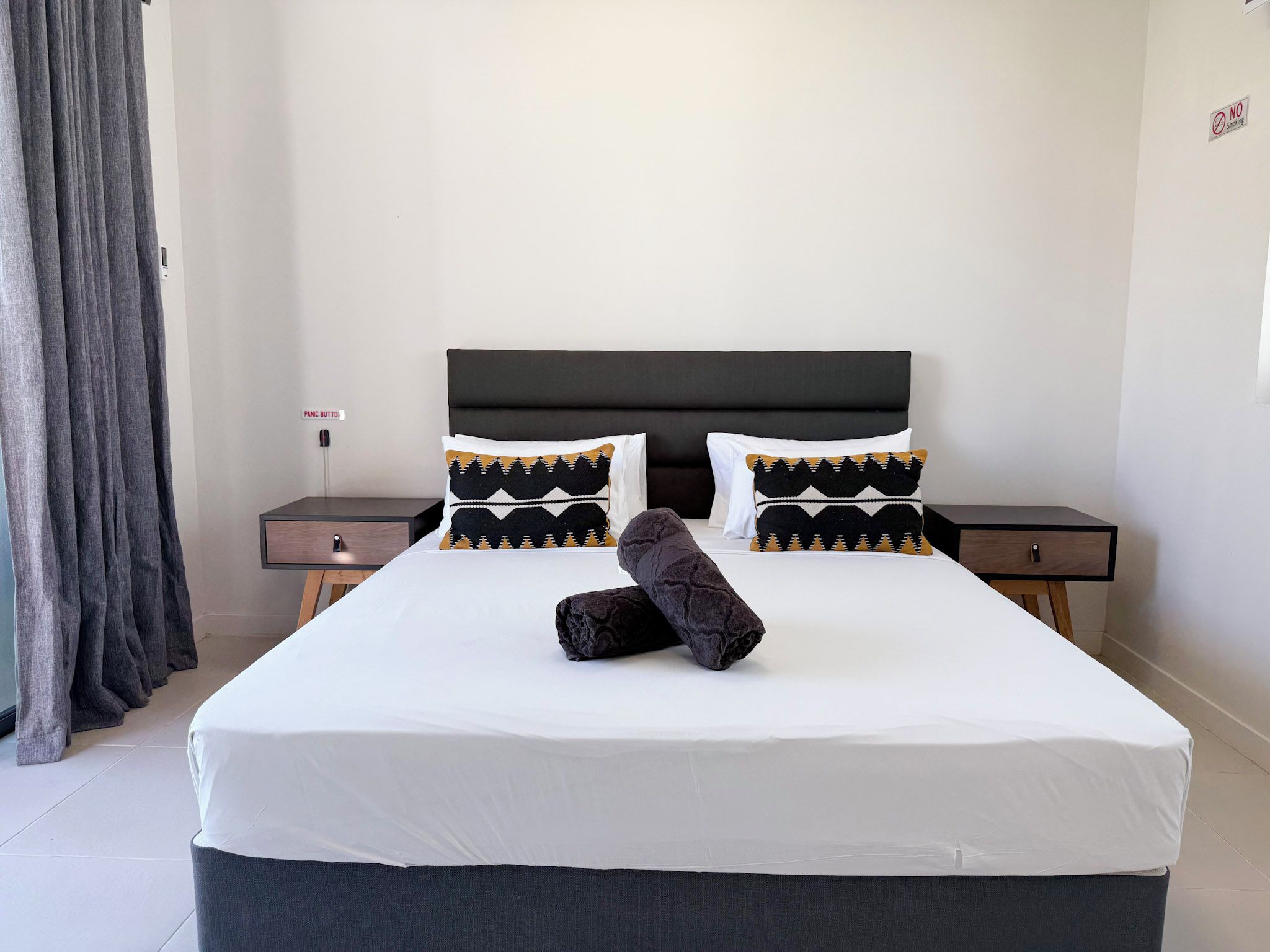 Una habitación de hotel con una cama grande, dos mesitas de noche, un cabecero y una cortina.