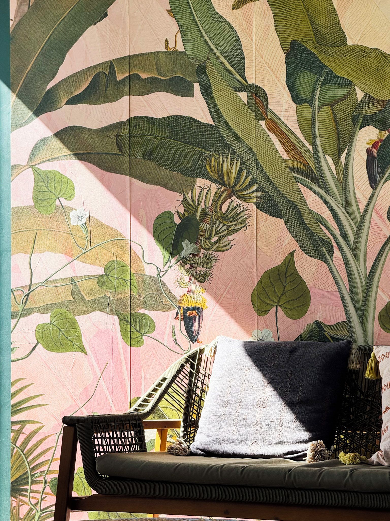 A room with a large botanical print on the wall, a wicker chair, and a cushion.